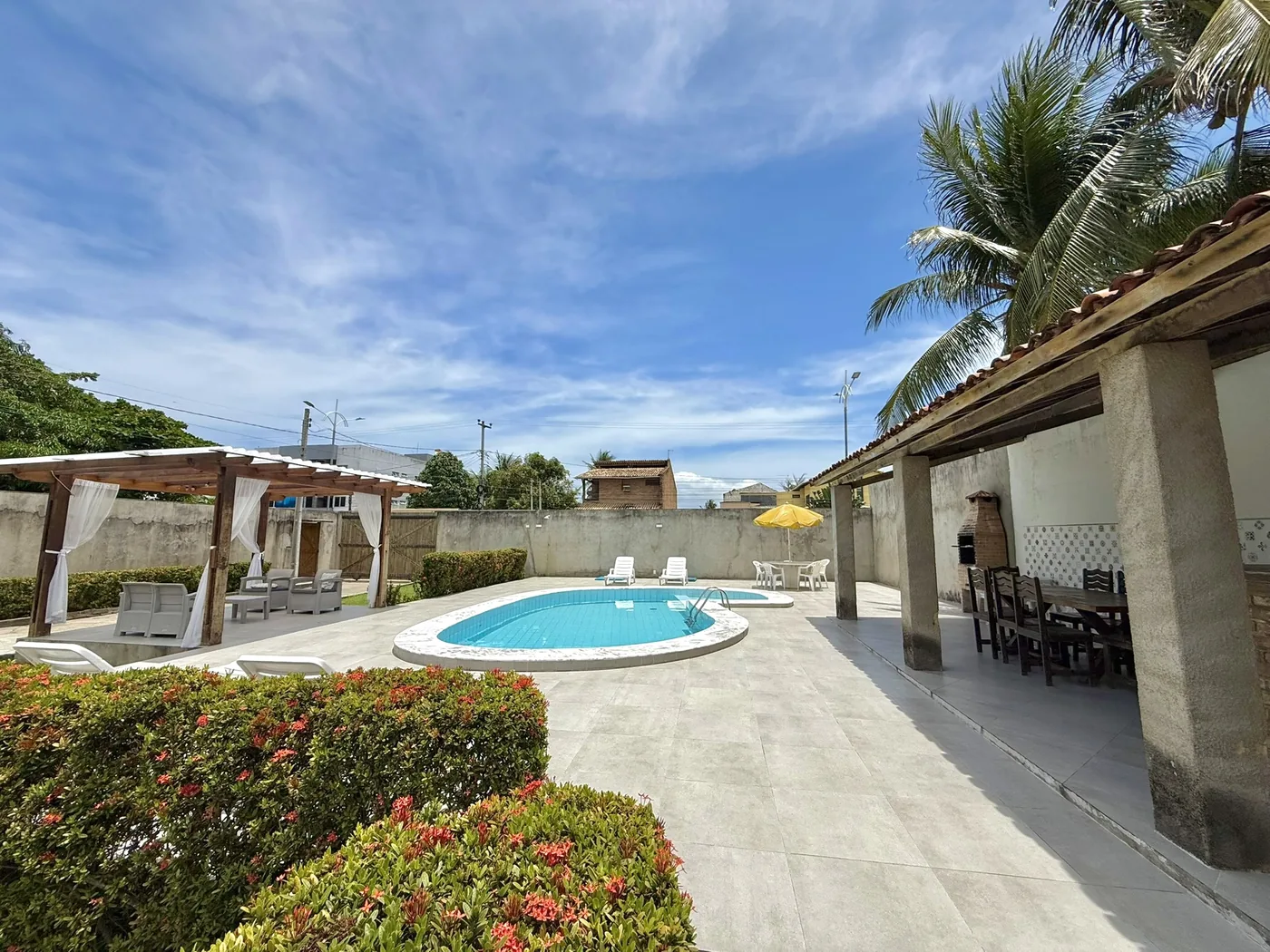 Piscina privativa da casa com pergolado de cortinas, espreguiçadeiras e área gourmet coberta com coqueiros