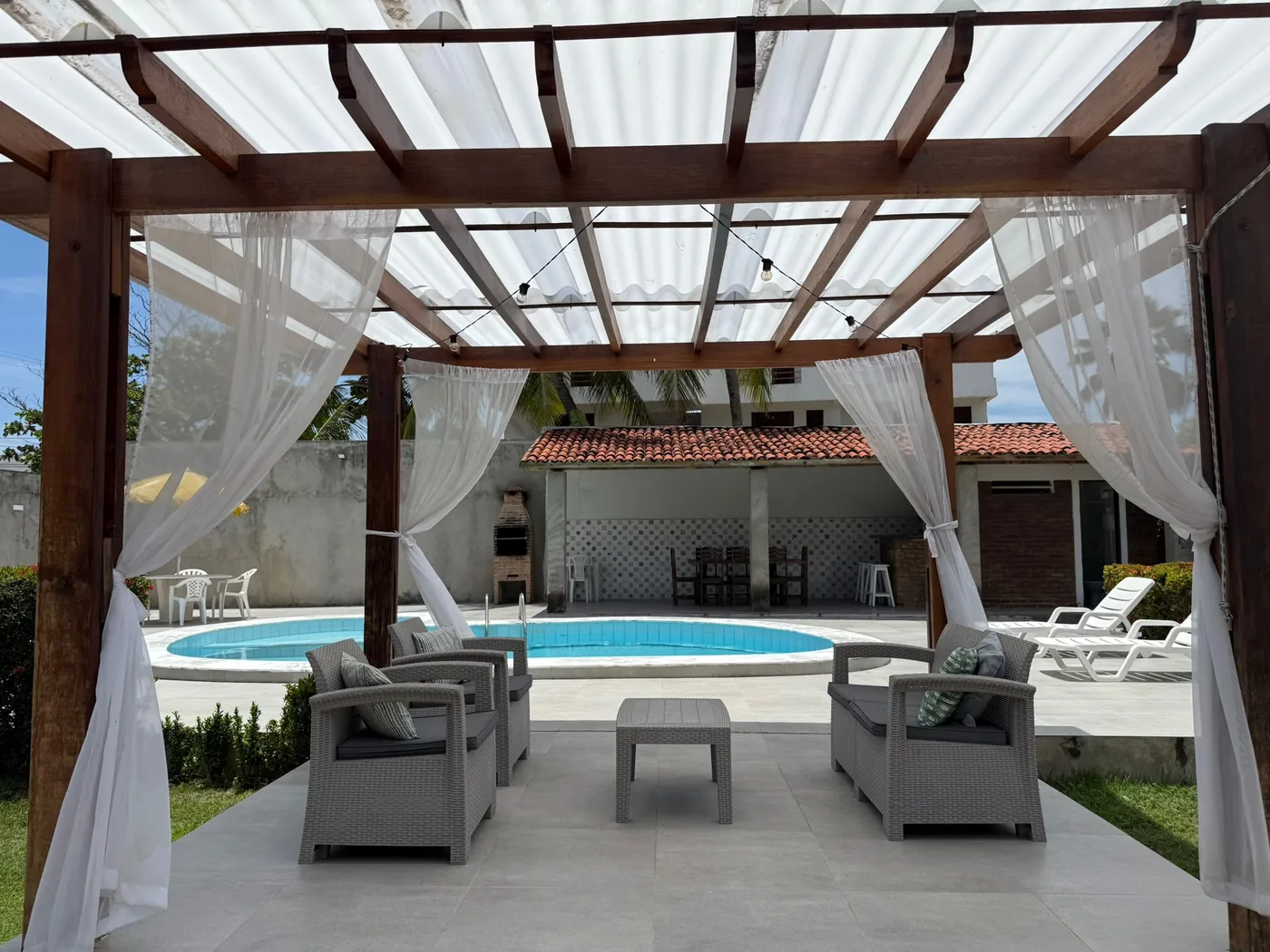 Pergolado com cortinas e poltronas de frente para a piscina